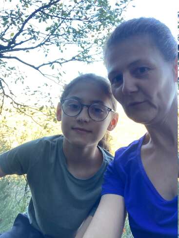 Eine Frau und ein Mädchen mit Brille stehen im Freien und posieren für ein Selfie. Sie sind von Bäumen und natürlichem Licht umgeben und sehen gemeinsam zufrieden und entspannt aus.