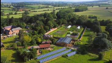 Esta imagen muestra una pintoresca granja rural con campos verdes, paneles solares, invernaderos, varios edificios, jardines y árboles, rodeada de campo y colinas lejanas.