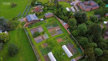 Esta imagen aérea muestra una exuberante y verde granja con múltiples hileras de cultivos, invernaderos, árboles circundantes, graneros y edificios cercanos, probablemente parte de una propiedad rural más grande.