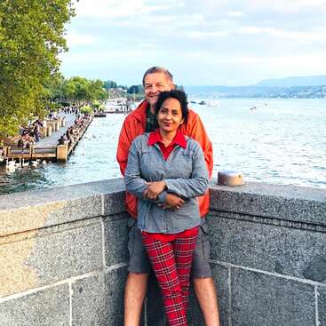 The image depicts a couple standing on a stone wall, with a serene lake and dock in the background, surrounded by lush greenery and a cloudy sky.