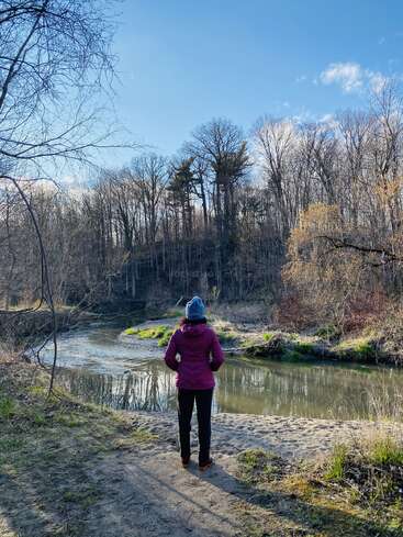 Eine Frau steht auf einem sandigen Weg neben einem Fluss, umgeben von Bäumen und Gras, und trägt eine rosa Jacke, eine schwarze Hose und einen blauen Hut.