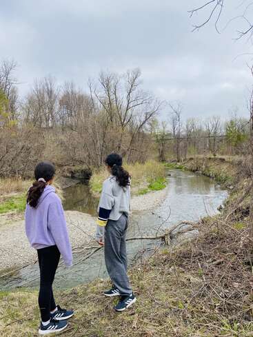 Zwei Frauen stehen am Ufer eines Flusses, umgeben von Bäumen und Gras, mit einem wolkenverhangenen Himmel über sich, und blicken auf das Wasser hinaus.