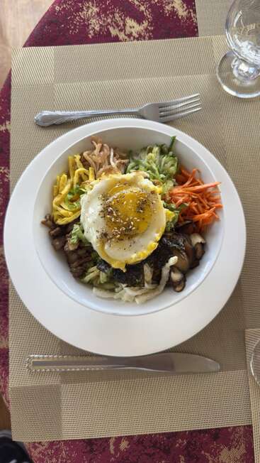 A beautifully arranged bowl featuring a sunny-side-up egg, sliced vegetables, mushrooms, seaweed, and meat, set on a placemat with utensils and a glass.