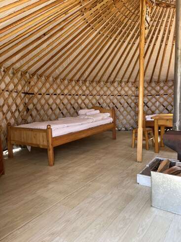 This image shows the inside of a cozy yurt with a wooden bed, small table and chairs, wooden floor, and a fireplace with firewood.