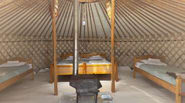 This image shows the cozy interior of a yurt with three wooden beds, each neatly made. A central wood stove provides warmth; lattice walls surround the space.