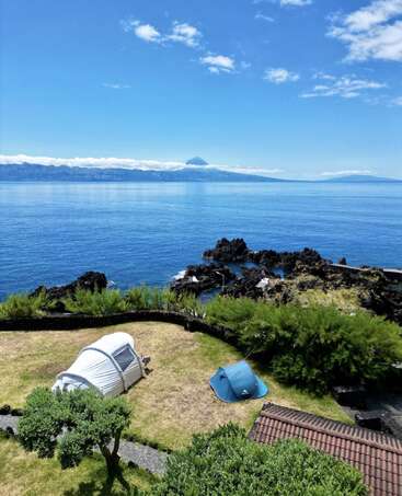 Deux tentes sont installées sur une pelouse au-dessus de roches volcaniques noires, surplombant les eaux calmes et bleues de l'océan et une montagne lointaine partiellement recouverte de nuages sous un ciel bleu éclatant.