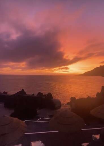 Un magnifique coucher de soleil illumine le ciel de teintes vives orange, roses et violettes, sur fond d'océan calme, de rochers se découpant sur la ligne d'horizon et de parasols de plage au premier plan.
