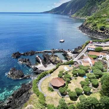 Cette image montre un magnifique paysage côtier avec une eau bleue cristalline, des côtes rocheuses, un petit port de plaisance, des jardins verdoyants, des bâtiments éparpillés et des montagnes luxuriantes sous un ciel clair.