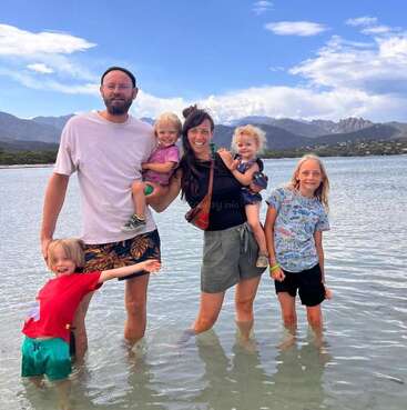 A family of five stands smiling in shallow water at the beach. Mountains and clouds are visible in the background. The children appear joyful and playful.