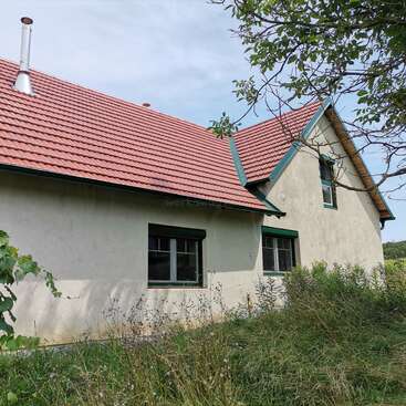 A quaint house with red-tiled roof, cream-colored walls, green window frames, and overgrown grass in front, surrounded by some trees under a clear sky.