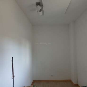 This image shows a small, empty room with white walls, a wooden floor, a single ceiling light, and a stick or mop leaning against the wall.