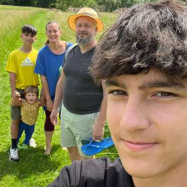 Eine fünfköpfige Familie posiert für ein fröhliches Gruppen-Selfie im Freien, lächelnd an einem sonnigen Tag auf einer Wiese mit Bäumen im Hintergrund.