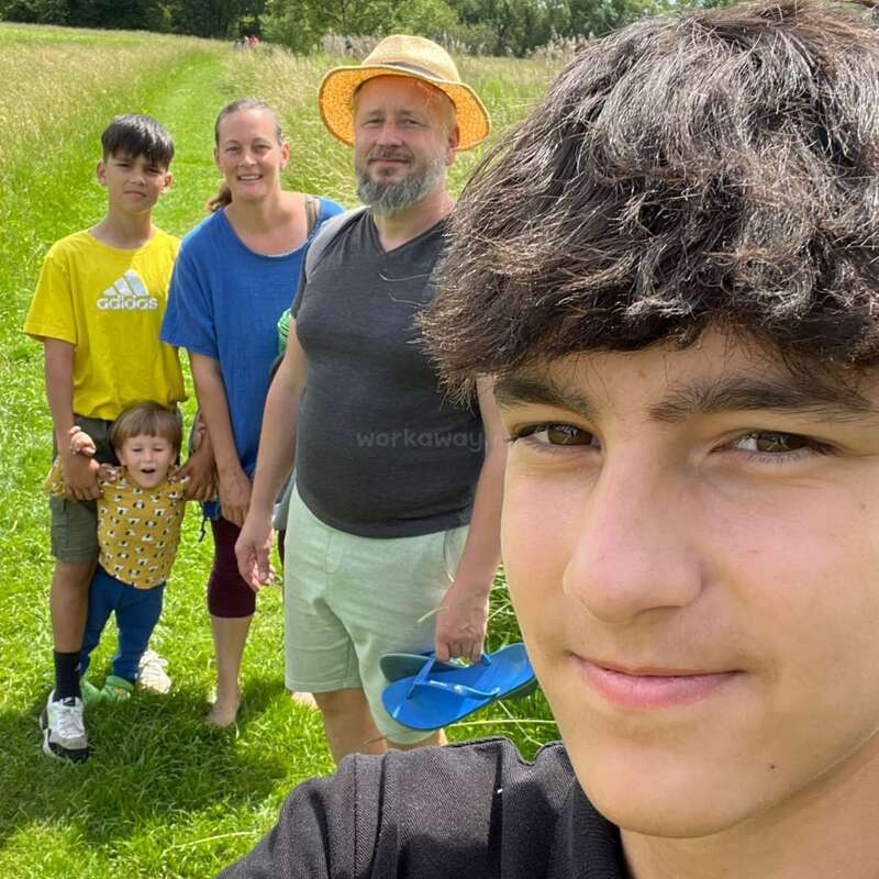 A family of five poses for a cheerful group selfie outdoors, smiling on a sunny day in a grassy field with trees in the background.