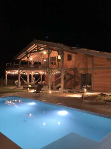 Une maison en bois confortable brille chaleureusement la nuit, avec ses guirlandes lumineuses et ses lanternes. Une piscine illuminée et sereine reflète l'ambiance accueillante de l'espace extérieur.