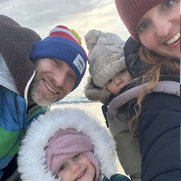 Une famille de quatre personnes, vêtue chaudement de bonnets et de manteaux d'hiver, sourit ensemble à l'extérieur par une journée froide, avec la plage et l'océan en arrière-plan.