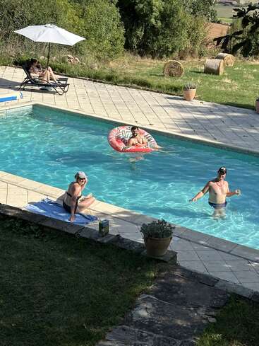 Quatro pessoas estão relaxando à beira da piscina: uma em uma espreguiçadeira sob um guarda-sol, outra em uma boia, outra sentada à beira da piscina e outra em pé na água.
