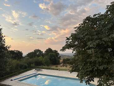Uma cena serena no campo mostra uma piscina cristalina, vegetação exuberante, espreguiçadeiras, fardos de feno ao longe e um céu colorido e parcialmente nublado ao pôr do sol.