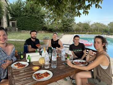 Cinco amigos estão sentados ao redor de uma mesa de madeira, saboreando pizza e bebidas ao ar livre, à beira da piscina, com árvores e vegetação ao fundo, sorrindo.