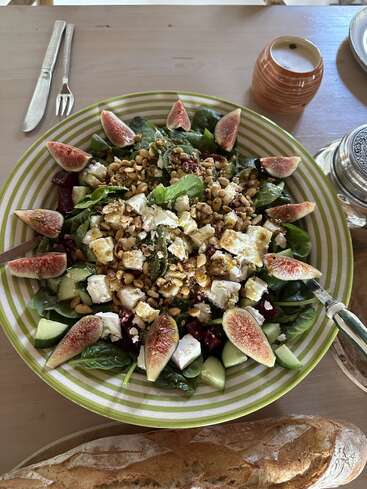 Uma salada colorida com figos frescos, espinafre, queijo feta, pepino, pinhões e, talvez, beterraba, servida numa tigela grande listrada, acompanhada de pão crocante.