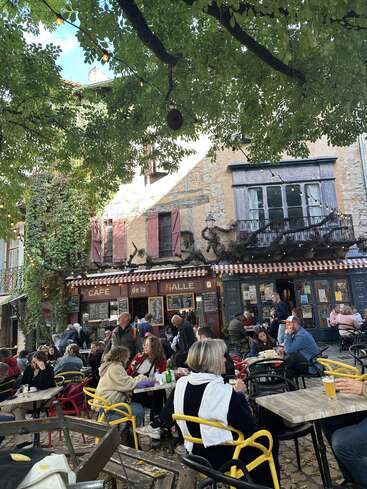 As pessoas estão tomando uns drinques e batendo papo no café ao ar livre “Café de la Halle”, sob árvores frondosas, com um charmoso prédio antigo de pedra ao fundo.