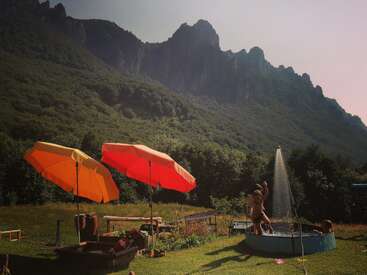 Des enfants jouent dans une petite piscine sous le soleil, près de parasols brillants, entourés d'arbres verts et de montagnes spectaculaires en arrière-plan. Plaisirs d'été en plein air.