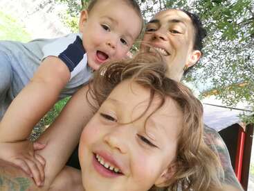 Un selfie joyeux montre un adulte souriant et deux enfants heureux à l'extérieur, sous un arbre. Le groupe rayonne de bonheur, avec des expressions enjouées et une lumière naturelle éclatante.