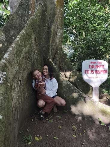 Une femme et un enfant sourient, assis au pied d'un grand chilamate (Ficus insipida) dans une forêt luxuriante et verdoyante. Panneau visible.