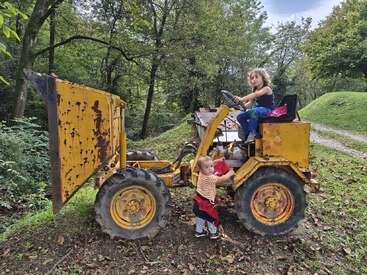 Deux enfants jouent sur un tracteur jaune rouillé dans une forêt verdoyante. L'un est assis au volant, tandis que l'autre se tient à côté, explorant les environs avec curiosité.