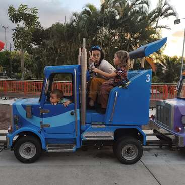 Une femme et deux enfants montent dans un petit camion bleu de parc d'attractions. La femme fait le signe de la paix tandis que les enfants semblent excités et captivés.