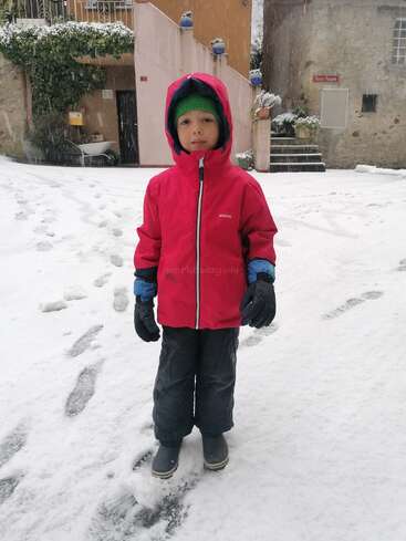 Un jeune enfant se tient debout dans la neige fraîche, vêtu d'une veste rouge, d'un bonnet vert, de gants noirs et de bottes. La neige recouvre le sol et les bâtiments environnants.
