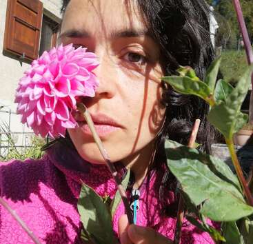 Une femme aux cheveux foncés tient une fleur rose vif devant son visage, couvrant partiellement un œil. La lumière du soleil et les ombres jouent sur ses traits.