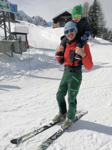 Un homme vêtu d'une tenue de ski verte porte un enfant sur ses épaules tout en skiant. Tous deux portent des lunettes de soleil et des bonnets d'hiver, entourés de neige et d'un paysage alpin.