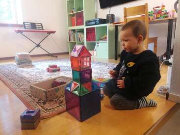 Un enfant en bas âge est assis par terre, occupé à construire une tour colorée avec des tuiles magnétiques. Des jouets, des étagères et un clavier remplissent cette pièce confortable et bien éclairée.