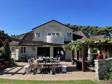 L'image représente une maison spacieuse à deux étages avec un patio, doté d'une table à manger et de chaises, entouré d'une végétation luxuriante et d'un ciel bleu serein.