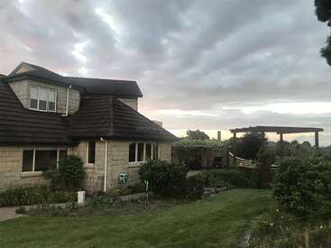 L'image représente une maison à deux étages au toit brun et à l'extérieur en pierre, entourée d'une végétation luxuriante et d'un ciel nuageux, dans un décor extérieur serein.