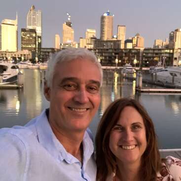 A imagem mostra um homem e uma mulher posando para uma selfie em frente ao horizonte de uma cidade e a uma marina, com os rostos do casal borrados.
