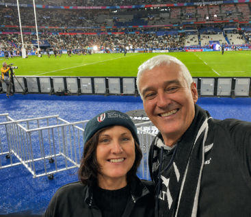 A smiling couple poses for a selfie at a rugby stadium during the Rugby World Cup 2023 in France, with a lively crowd in the background.