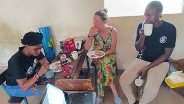 Three women sit around a small table in a casual room, eating snacks, drinking from mugs, and chatting. A laptop and food items are on the table.