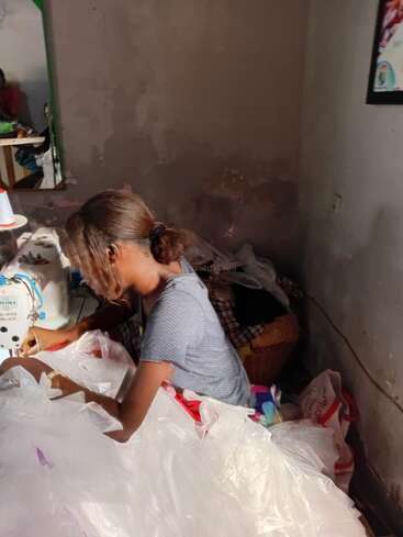 A woman sits at a sewing machine, working with white fabric or plastic material in a dimly lit room with worn walls and scattered items.