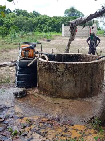 A concrete well with a water pump ejecting water onto the ground, surrounded by greenery, tires, a building in the background, and a person standing nearby.