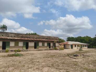 This image shows two old, simple school buildings with rusted roofs on dry, bare ground. The sky is partly cloudy, and some vegetation surrounds the area.