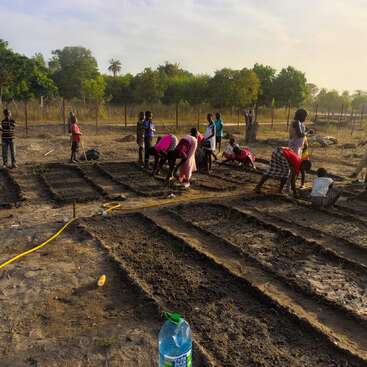 A group of people, including children, are working together on a garden, tending to soil beds. The setting sun lights a rural, green landscape.