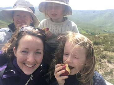 A smiling family of four enjoys a day outdoors on a sunny mountain hike. Two children are eating apples while the parents pose happily together.