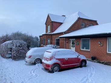 A red brick house with snow-covered roof stands in a snowy yard. Two cars parked outside are covered in snow. Bushes and “For Sale” sign visible.