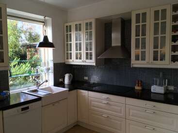 This kitchen is bright with large windows, white cabinets, black countertops, a farmhouse sink, modern appliances, a tiled backsplash, and glass-front cupboards. It's organized and inviting.