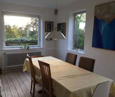 This dining room features a rectangular table with a yellow cloth, six chairs, large windows, potted plants, wall art, wooden floors, and a modern hanging lamp.