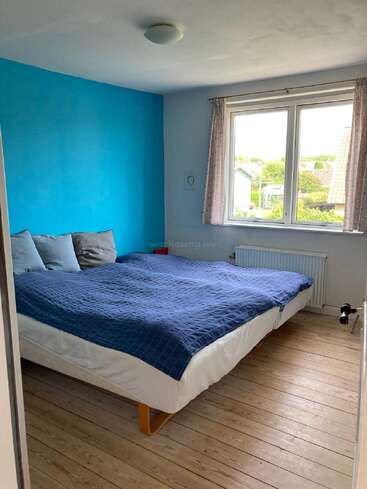 A cozy bedroom with wooden floors, a large bed with blue bedding, three gray pillows, a blue accent wall, window with curtains, and natural light.