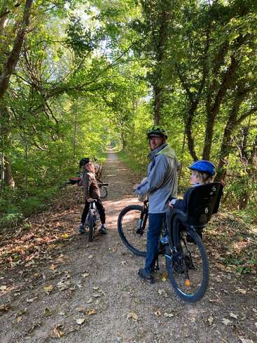 A man and two children, all wearing helmets, pause during a bike ride on a leafy, sunlit forest trail, enjoying nature and each other's company.