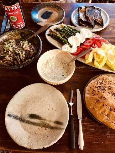 A rustic table features a spread of fresh bread, tzatziki, salad, grilled asparagus, roasted eggplant, onions, cheese, tomatoes, and a bottle of red wine.