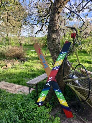 In a lush green garden, two colorful wooden signs lean against a tree. One reads "Vielfalt und bunt statt" and the other "umrja," surrounded by nature.